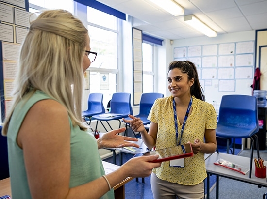 An over-the-shoulder view of two female teachers having a pre-school meeting and sharing teaching ideas with each other. They are going over the lesson plans and checking they are all prepared on their digital tablets.