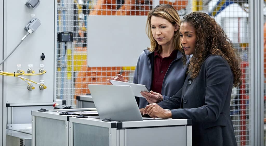 Two women dressed professionally on factory floor reviewing a tablet and laptop