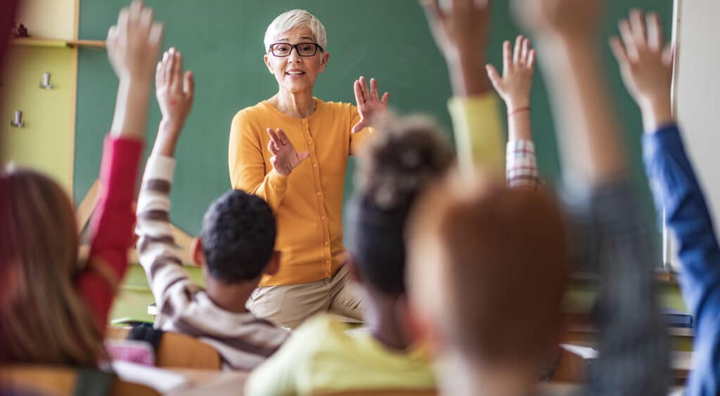 Photo of teach facing a room full of students with raised hands