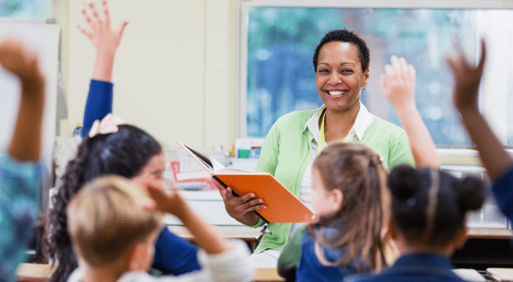 A teacher in a classroom with students raising their hands.